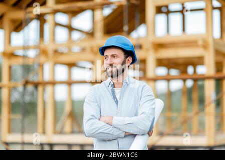 Portrait d'un architecte ou d'un constructeur en casque de sécurité debout devant la structure de la maison en bois. Construction et conception de maisons en bois Banque D'Images