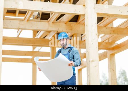 Builder en combinaison bleue et casque de chantier avec des plans sur le chantier. Construction en bois cadre maison concept Banque D'Images