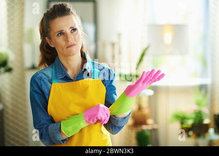 malheureux moderne 40 ans femme en tablier orange et des gants en caoutchouc rose dans la maison moderne en journée ensoleillée commencer les travaux ménagers. Banque D'Images