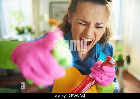 furieuse moderne 40 ans femme en tablier orange et des gants en caoutchouc rose avec un flacon pulvérisateur de fournitures de nettoyage menaçant avec le doigt dans le moderne Banque D'Images