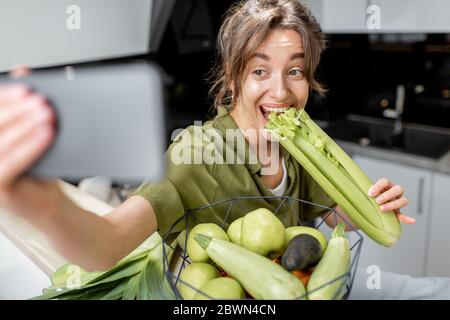 Jeune femme faisant selfie photo ou vlogging sur téléphone mobile au sujet de la saine alimentation, assis avec des ingrédients crus sur la cuisine à la maison. Concept de mode de vie sain et d'influence sociale Banque D'Images