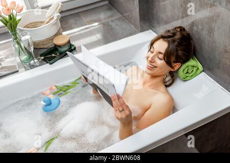 Jeune femme détendue prenant un bain, allongé dans la baignoire et lisant un magazine à la maison Banque D'Images