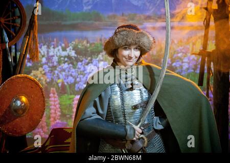 Une femme vêtue de courrier en chaîne, de manteau et de chapeau de fourrure avec un scimitar dans ses mains contre le fond de champ avec des fleurs, rivière et montagnes dans le Banque D'Images