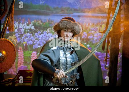 Une femme vêtue de courrier en chaîne, de manteau et de chapeau de fourrure avec un scimitar dans ses mains contre le fond de champ avec des fleurs, rivière et montagnes dans le Banque D'Images
