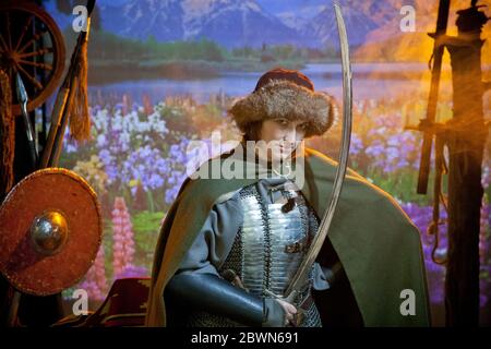 Une femme vêtue de courrier en chaîne, de manteau et de chapeau de fourrure avec un scimitar dans ses mains contre le fond de champ avec des fleurs, rivière et montagnes dans le Banque D'Images