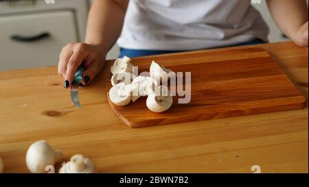 Jeune femme coupant des champignons sur bois dans la cuisine. Coupe féminine de champaignon blanc avec couteau sur planche à découper sur table de cuisine. Banque D'Images