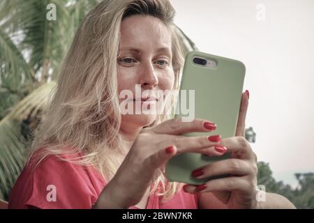Femme blonde adulte tenant, utilisant et regardant le smartphone extérieur Banque D'Images