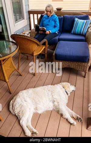 Femme âgée lisant un livre sur un patio résidentiel extérieur avec un chien Golden Retriever de couleur platine Banque D'Images