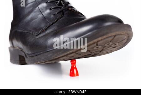 Chaussure noire de classe qui fait des stomères et écrasent un petit morceau de jeu de pion rouge sous la semelle. Grand vs petit, pouvoir, danger et impuissance concept abstrait Banque D'Images