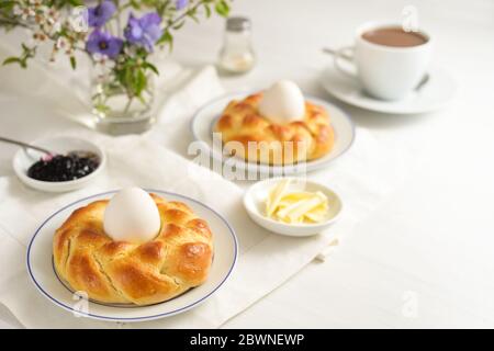 Pain de levure de Pâques plaquée en nid avec des œufs, du café, du beurre, de la confiture et des fleurs sur une table blanche, comme un petit déjeuner de dimanche festif, espace de copie, sélectionnez Banque D'Images