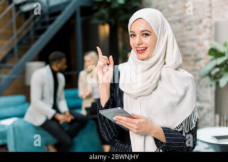 Jolie jeune femme d'affaires musulmane en hijab blanc debout dans la salle de bureau moderne, tenant un PC tablette numérique, souriant avec son doigt pointant Banque D'Images