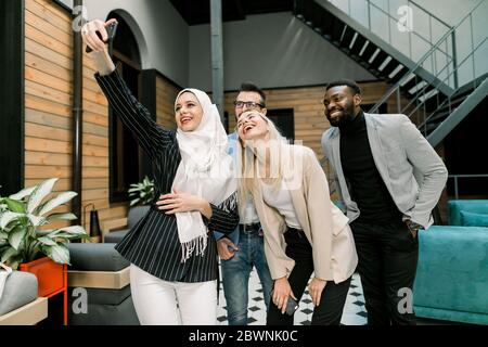 Joyeux quatre jeunes gens d'affaires multiethniques attrayants prenant photo selfie après un travail ou un projet réussi, ou appréciant leur temps libre de déjeuner à l'intérieur Banque D'Images