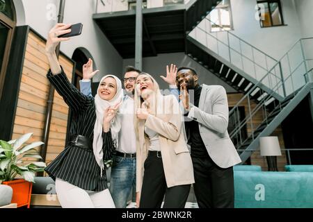 Souriant quatre jeunes collègues multiethniques, ils font des selfies ensemble, sourient et montrent des gestes différents à la main à l'appareil photo Banque D'Images