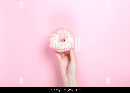 La main de femme tient le donut avec glaçage rose et garniture de pâtisserie de noix de coco, fond rose Banque D'Images