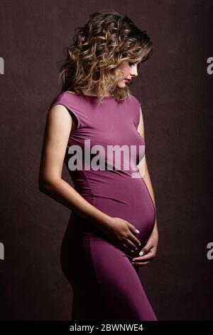 Une belle femme enceinte met ses mains sur son ventre et le regarde. Femme avec coiffure et maquillage. Photo prise dans un studio de photo sur un b marron Banque D'Images