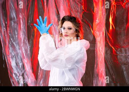 Jeune fille médecin en lunettes met sur des gants, fond de cellophane avec lumière rouge. Photographie d'art Banque D'Images