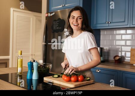 Belle jeune femme brune cuisant de la salade en se tenant au comptoir de la cuisine Banque D'Images