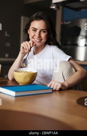 Belle jeune femme souriante brune ayant un petit déjeuner sain tout en étant assise à la table de cuisine Banque D'Images