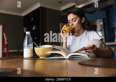 Jeune femme brune attrayante ayant un petit déjeuner sain lisant un livre tout en étant assis à la table de cuisine Banque D'Images