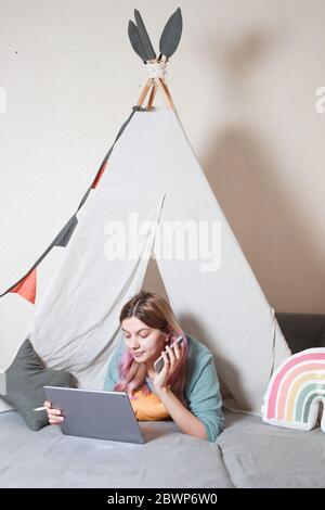 Une jeune mère travaille sur une tablette à la maison, dans une cabane pour enfants faite de draps. Concept de quarantaine pendant une pandémie Banque D'Images