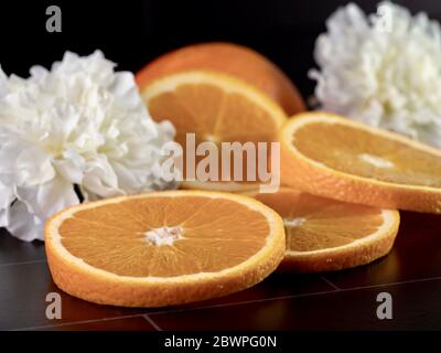 Tranches d'orange frais et une demi-orange sur un comptoir de carreaux noirs avec deux œillets blancs pour la décoration. Agrumes frais photo avec sélectiv Banque D'Images