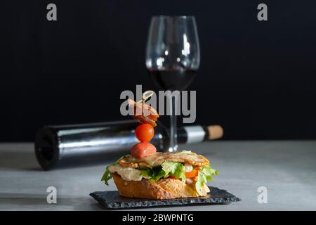 Gros plan d'un délicieux sandwich ouvert avec du fromage et un assortiment de viandes et de légumes sur une assiette sombre avec un verre de vin dépoli Banque D'Images