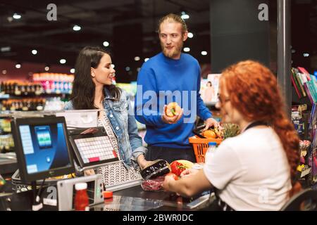 Un jeune couple souriant se tient près du comptoir de caisse tout en achetant des produits ensemble dans un supermarché moderne Banque D'Images