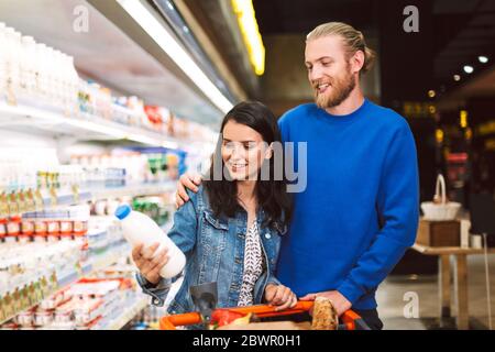 Jeune couple souriant avec chariot plein de produits heureux de choisir le lait dans le supermarché Banque D'Images