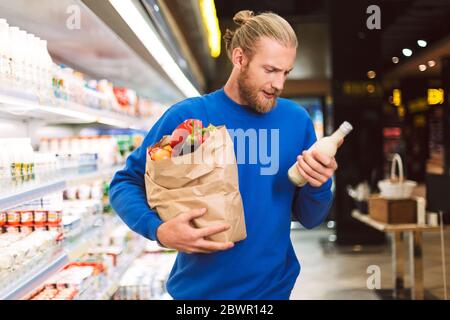 Jeune homme en chandail bleu foncé tenant le sac d'épicerie en papier plein de produits tout en choisissant soigneusement le lait dans le département de lait du supermarché moderne Banque D'Images