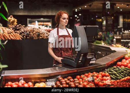 Jeune vendeur en tablier derrière le comptoir avec des légumes travaillant dans un supermarché moderne Banque D'Images