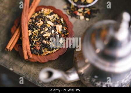 Feuilles aromatiques fruitées de thé noir séchées et bandes d'écorce de cannelle posées sur un petit plateau en bois avec une bouilloire métallique au premier plan Banque D'Images