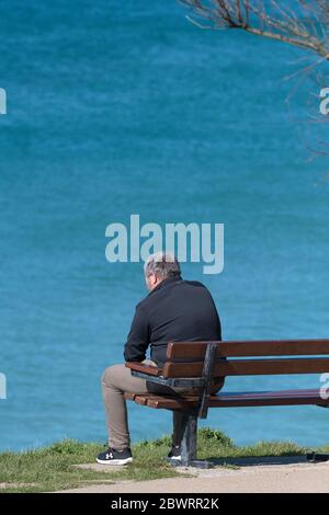 Un homme assis seul sur un banc surplombant la mer à Newquay, en Cornouailles. Banque D'Images