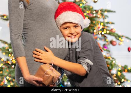 Portrait d'un petit garçon portant un chapeau de père Noël câlin le ventre de la mère tenant présent debout près de l'arbre du nouvel an Banque D'Images