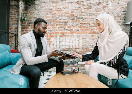 Photo de bureau d'une femme d'affaires arabe musulmane souriante satisfaite des résultats des négociations, signant un accord avec son beau homme africain Banque D'Images