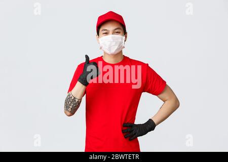 Ne vous inquiétez pas de votre colis dans nos mains. Homme de livraison asiatique souriant et confiant, uniforme rouge, masque médical et gants de protection, pouces-up dedans Banque D'Images