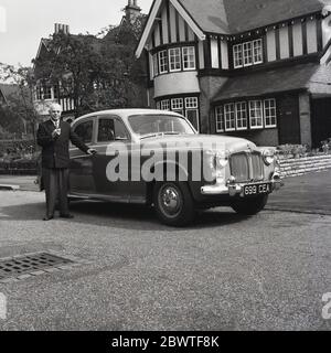 1961, historique, un vieil Anglais gentleman dans un balzer et des Noirs et tenant un tuyau, debout fièrement par son bicolore Rover 100 hors d'une grande maison dans la banlieue, Angleterre, Royaume-Uni. Conçues par Gordon Bashford et de la famille de véhicules Rover P4, connues sous le nom de « Auntie Rovers », ces élégantes voitures ont été pilotées par Royalty, propriété de Grace Kelly, et constituent une part importante de la culture britannique. Le Rover 100 a été lancé en octobre 1959 et était une berline 4 portes avec un intérieur luxueux en cuir et en bois. Banque D'Images