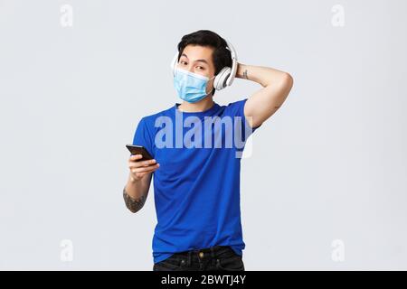 Covid-19 mode de vie, émotions et loisirs sur le concept de quarantaine. Beau jeune homme asiatique queer dans le masque médical et le casque, écouter de la musique Banque D'Images