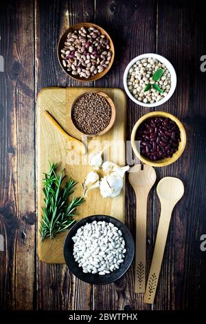 Planche à découper, louches et cuillère en bois, ail, romarin et bols avec divers haricots et lentilles Banque D'Images