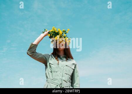 Jeune femme à tête rouge contre le ciel couvrant les yeux avec un bouquet de fleurs jaunes Banque D'Images