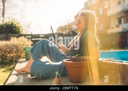 Jeune femme dans le jardin avec semis dans le pot de fleurs à l'aide d'un smartphone Banque D'Images