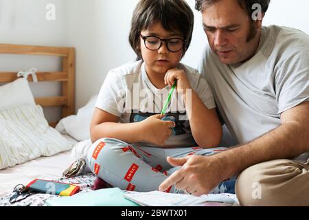 Père assistant son fils dans les devoirs pendant qu'il est assis sur le lit à la maison Banque D'Images