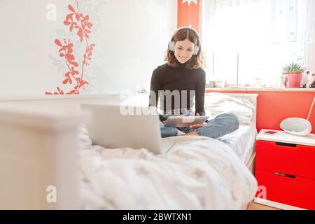 Portrait d'une adolescente souriante avec casque assis sur le lit à l'aide d'un ordinateur portable et d'une tablette numérique Banque D'Images