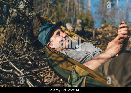 Jeune homme se détendant dans un hamac, en utilisant un smartphone Banque D'Images