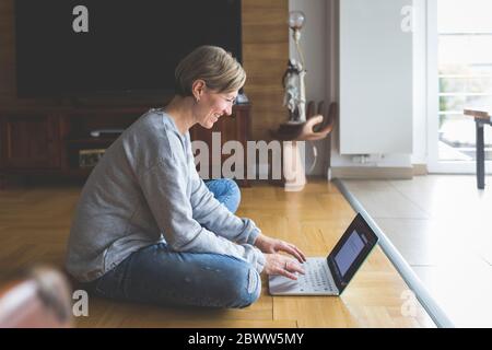 Femme mature travaillant à la maison, utilisant un ordinateur portable, assise à pieds croisés sur le sol Banque D'Images