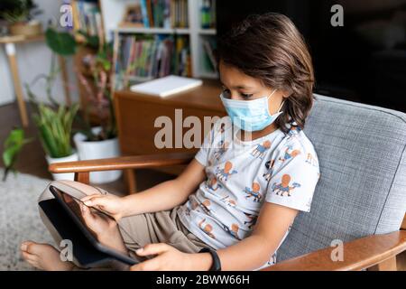 Garçon avec masque chirurgical assis sur un fauteuil à la maison en utilisant une tablette numérique Banque D'Images