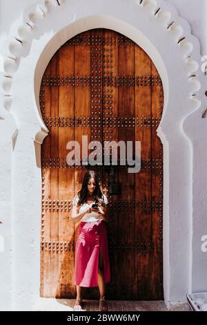 Jeune femme debout devant la porte d'entrée traditionnelle regardant le téléphone cellulaire, Merzouga, Maroc Banque D'Images