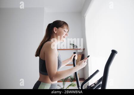 Femme utilisant un smartphone tout en effectuant un entraînement sur vélo elliptique à la maison Banque D'Images