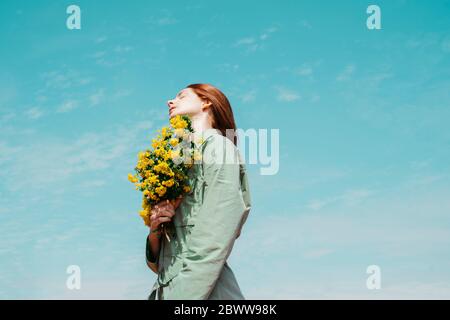 Jeune femme à tête rouge avec les yeux fermés debout contre le ciel tenant un bouquet de fleurs jaunes Banque D'Images