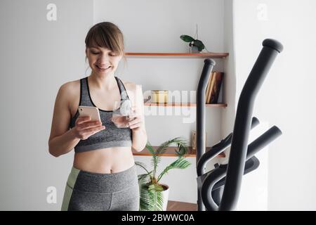 Femme buvant de l'eau et utilisant un smartphone après avoir exécuté l'entraînement sur l'entraîneur elliptique à la maison Banque D'Images