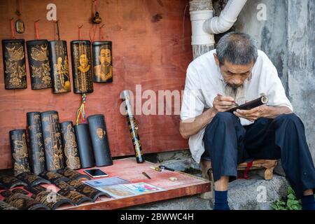 Feng Huang, Chine - août 2019 : ancien artiste chinois, homme assis sur le trottoir de la rue et se concentrant sur la réalisation d'une photo dessinée sur un bambou avec du penc Banque D'Images